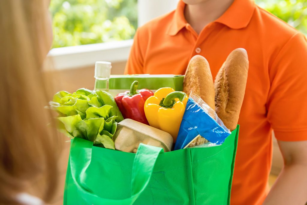 A woman with medium blond hair opens the door to a grocery delivery man in an orange polo shirt holding a green shopping bag filled with groceries
